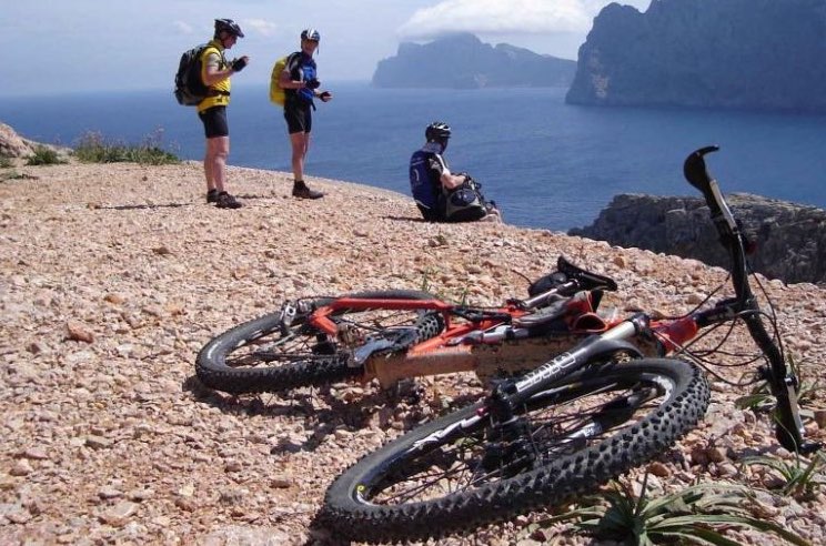 Mountainbike liegt auf Schotterweg und im Hintergrund blicken Fahrradfahrer aufs Meer.